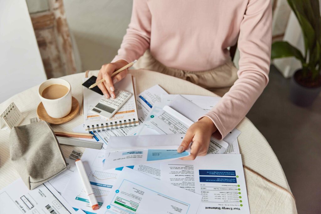 Cropped shot of unrecognizable woman calculates goods service price paying bills sits at round table with paper documents and coffee engaged in home banking manages finances. Financial paperwork